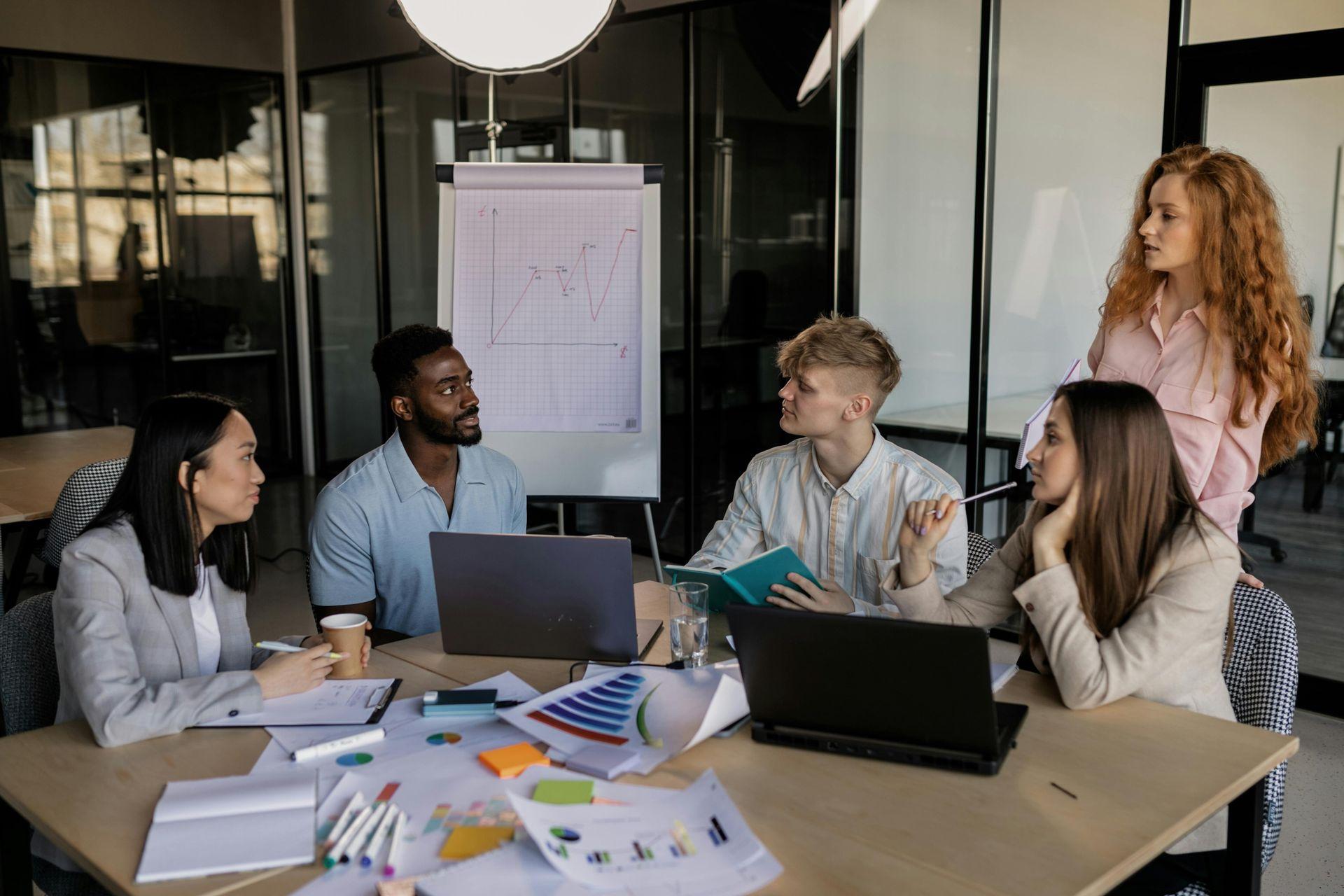 "A diverse team of business professionals engaged in a strategic consulting session, discussing business growth, financial planning, and nonprofit development. The modern office setting includes laptops, charts, and a flip chart displaying a rising graph, emphasizing collaboration, startup consulting, and business strategy planning."