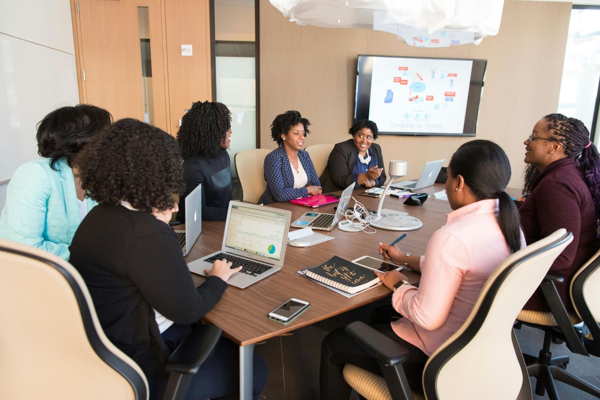 "A diverse group of professional women engaged in a boardroom meeting, collaborating around a large conference table with laptops, notebooks, and documents. A presentation screen in the background displays a business strategy discussion. The setting is a modern office environment, fostering teamwork and strategic planning."